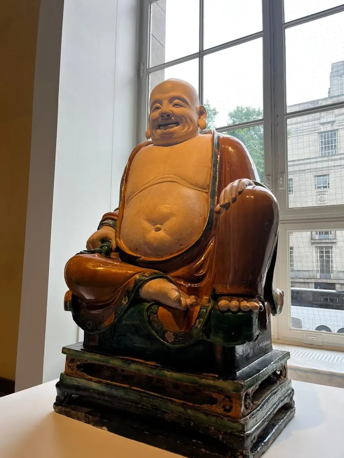 A laughing Buddha figure in glazed ceramic, lit by window light in a museum gallery