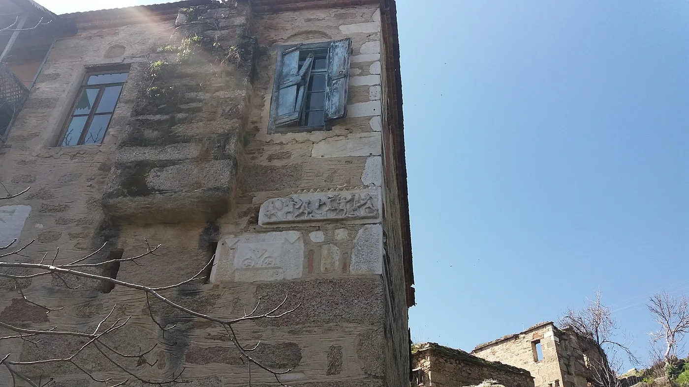 The facade of an old stone house built with reused ancient blocks, with wooden shutters and carved marble reliefs